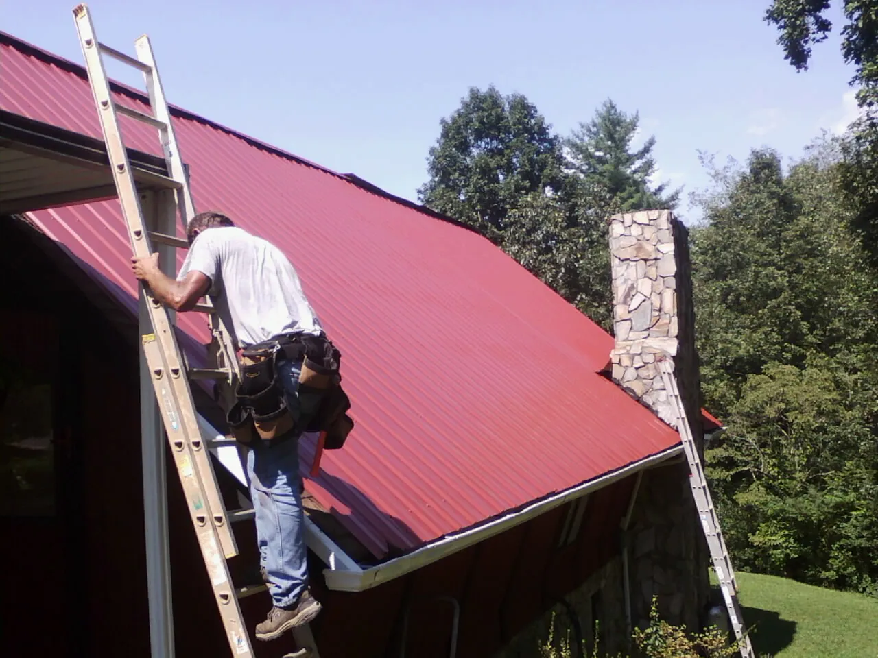 Roofer on a ladder beside a red metal roof and stone chimney — Soffit & Fascia Repair by Roofing Brothers in Seville